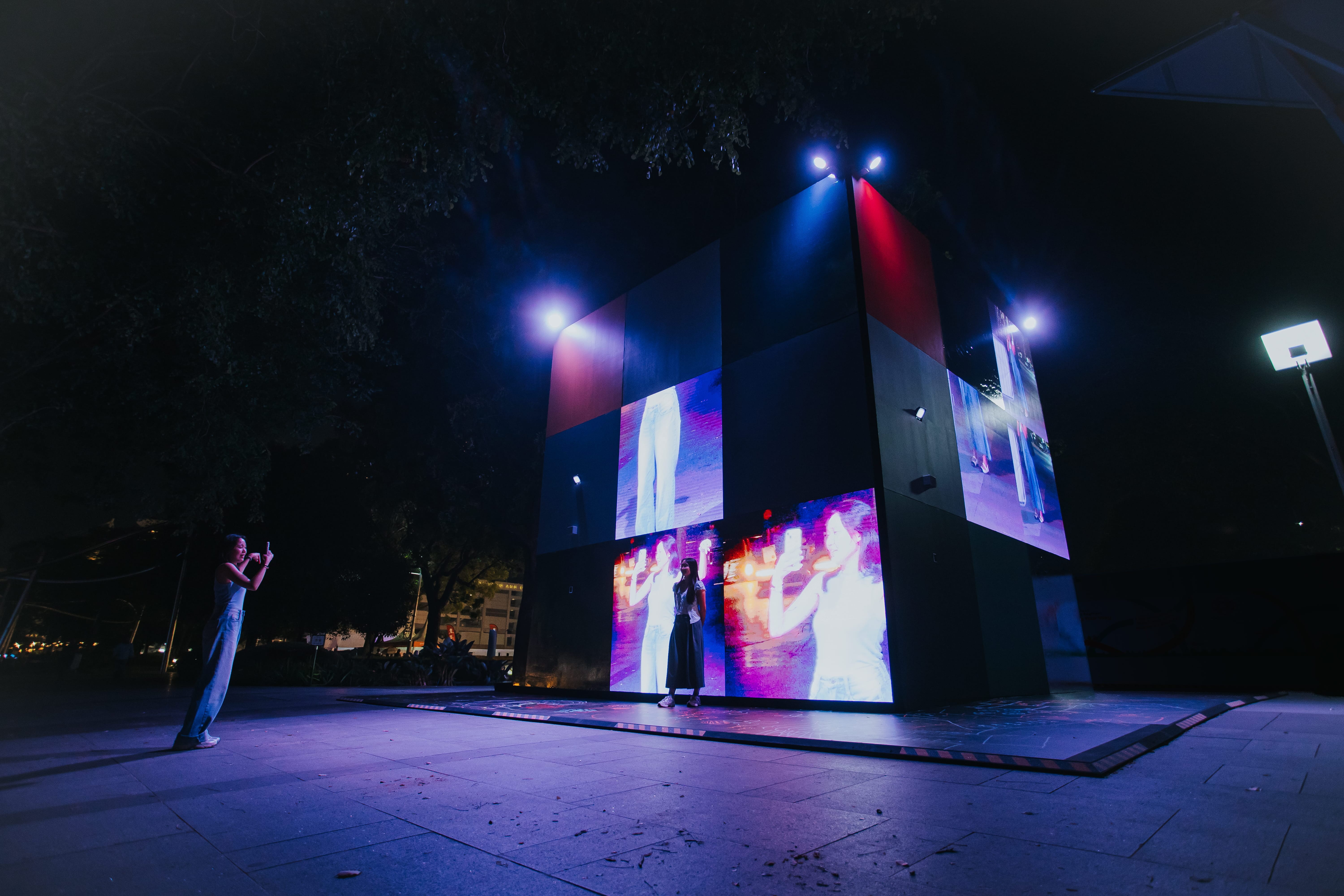 A cube-shaped public art installation at night, its panels showing fragmented facial close-ups and solid color blocks, with people gathered around it.